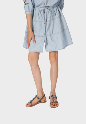 Donna che indossa un vestito azzurro chiaro con bottoni e cintura in vita e sandali bassi blu con decorazioni floreali gioiello, in piedi su uno sfondo semplice.