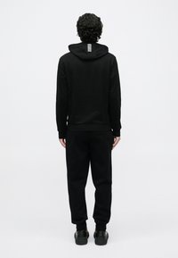 Persona con cabello rizado de espaldas vistiendo sudadera con capucha negra y pantalones deportivos con zapatillas negras sobre un fondo gris liso.