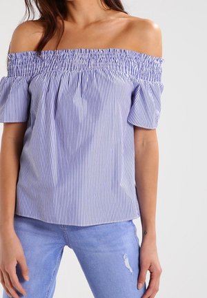 Kvinde iført en blå og hvid verticallystribet off-shoulder top kombineret med lyseblå, revnede jeans, stående mod en ensfarvet baggrund.