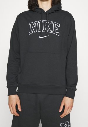 Osoba ubrana w czarną bluzę z kapturem Nike i dopasowane czarne spodnie dresowe, z białym logo i napisem Nike na bluzie i spodniach.