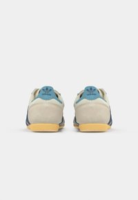 Pari di sneaker Adidas in suede beige, con accenti blu, suole in gomma e logo Adidas sulla linguetta del tallone, mostrati da dietro su sfondo bianco.