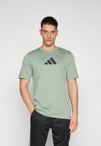 Zelené tričko s krátkým rukávem vyrobené z měkké látky, na přední straně s černým logem Adidas. Jednoduchý kulatý výstřih a volný střih.