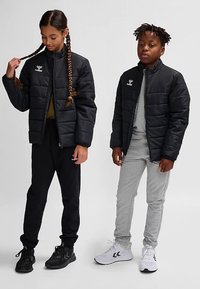 Sorte polstrede jakker med høje kraver, lynlås foran og logo; båret med grå og sorte atletikbukser og sneakers. Børn stående side om side.