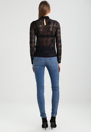 Femme portant un haut en dentelle noire à manches longues et un jean skinny bleu avec des boutons sur les côtés, debout de dos sur fond blanc.