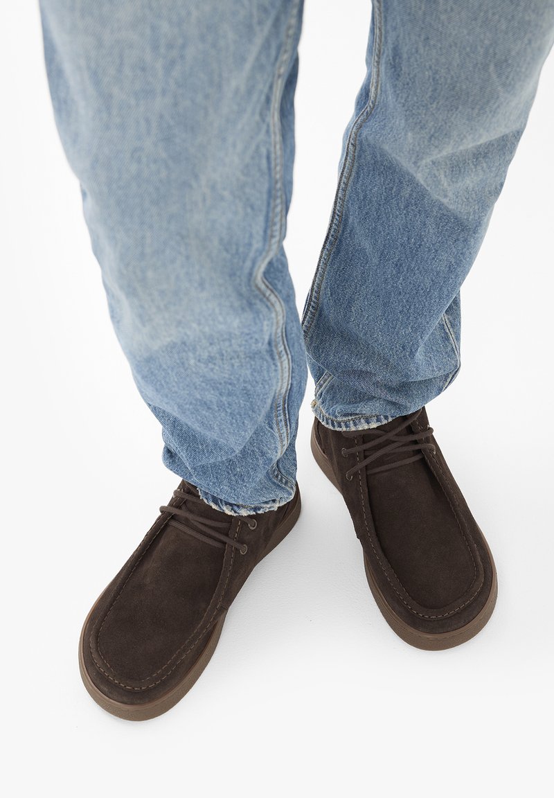 Personne portant un jean bleu clair et des chaussures en daim marron foncé à lacets, debout sur une surface blanche.