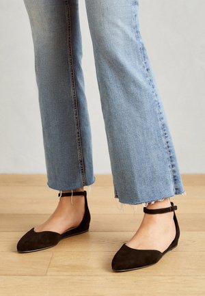 Black suede flats with a pointed toe and ankle strap, paired with cropped light blue jeans featuring frayed hems on a wooden floor.