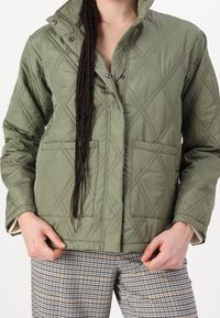 Quiltad, olivgrön jacka med hög krage, tryckknappar och två framfickor. Har ett diamantmönster och texturerad tyg.