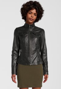 Zwarte leren jas met een hoge kraag, een getailleerd ontwerp, subtiele stikseldetails en twee voorkruisjes, gedragen over een geribde olijfgroene rok.