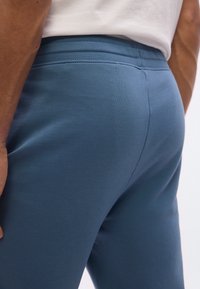 Κοντινό πλάνο ατόμου που φοράει μπλε εφαρμοστά παντελόνια και λευκό πουκάμισο, εμφανίζοντας τη μέση και την άνω περιοχή των μηρών από πίσω.