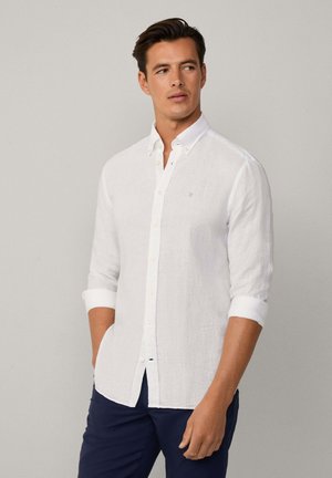 Camisa de lino blanca de botón con mangas arremangadas, que presenta un emblema azul claro en el pecho y botones en contraste. Textura suave.