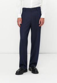 Pantalon formel bleu marine avec une coupe droite, devant plissé, passants de ceinture et fermeture à bouton. Le tissu semble lisse et structuré.