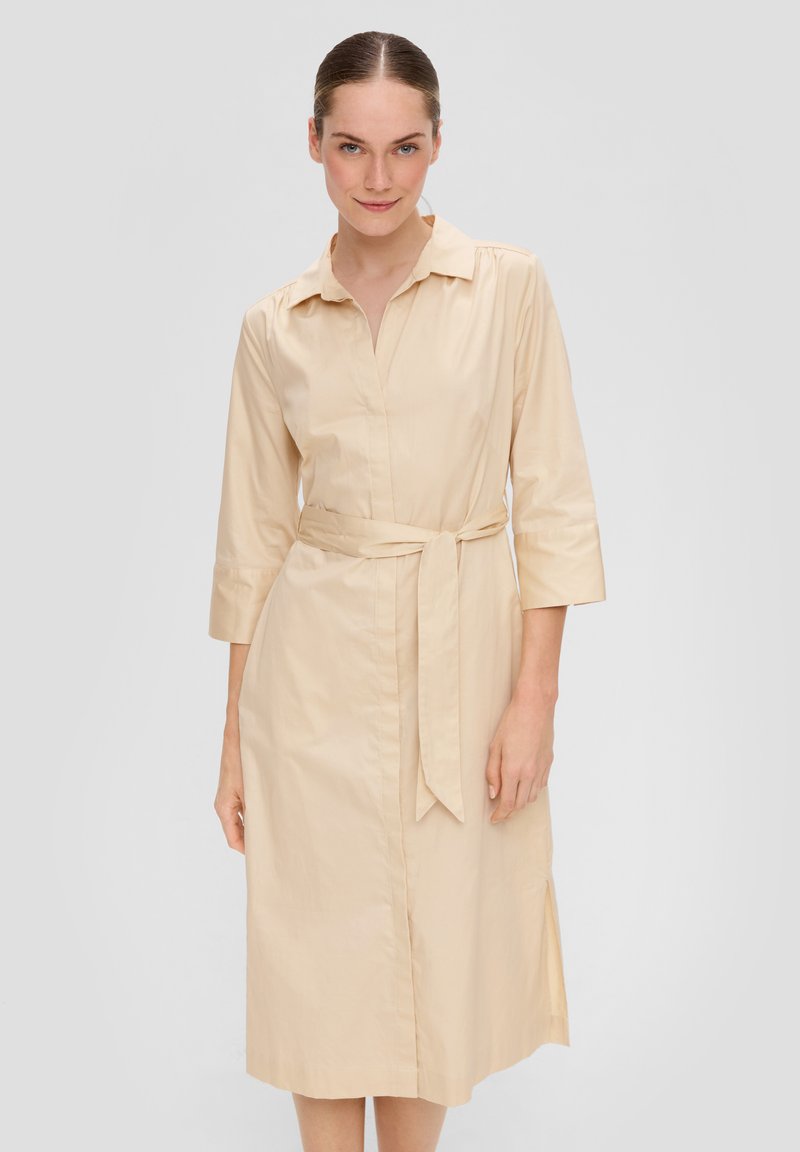Hellbeige Kleid mit Kragen, dreiviertel Ärmeln, tailliertem Design und geradem Saum. Sanfter Stoff mit seitlichen Schlitzen für mehr Bewegungsfreiheit.