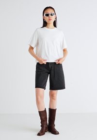T-shirt blanc à manches courtes, shorts en denim noir, et bottes marron à hauteur de genoux avec boucles. Accessoires comprenant des lunettes de soleil noires élégantes.