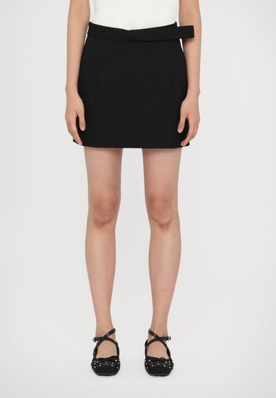 Mini-jupe noire au design ajusté, dotée d'une ceinture à la taille, de coutures discrètes et d'une texture lisse. Associée à des ballerines noires.