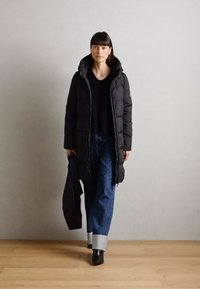 Black puffer coat with a hood and long sleeves, worn over a black top. Features a long, oversized shape and relaxed blue denim jeans.