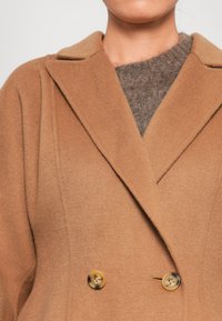 Manteau en laine couleur chameau avec un design à double boutonnage, présentant de grands boutons ronds et des revers crantés, superposé à un pull gris.