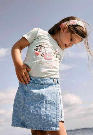 Jong meisje draagt een lichtblauw grafisch t-shirt en een blauwe spijkerrok met hartjespatroon, kijkt naar beneden met een bloemenhaarbandje in haar haar.