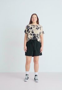 Top beige de manga corta con estampado floral negro, combinado con pantalones cortos negros de tiro alto con detalle de lazo y zapatillas negras.
