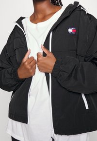 Svart vindjacka med elastiska ärmslut och huva, med en vit dragkedja och en liten färgad logotyp på bröstet. Bärs över en vit skjorta.