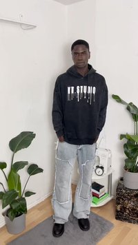 Camisola preta com o logotipo "MT STUDIO" em design branco escorrendo, combinada com jeans claros desgastados com remendos e rasgos, e botas pretas.