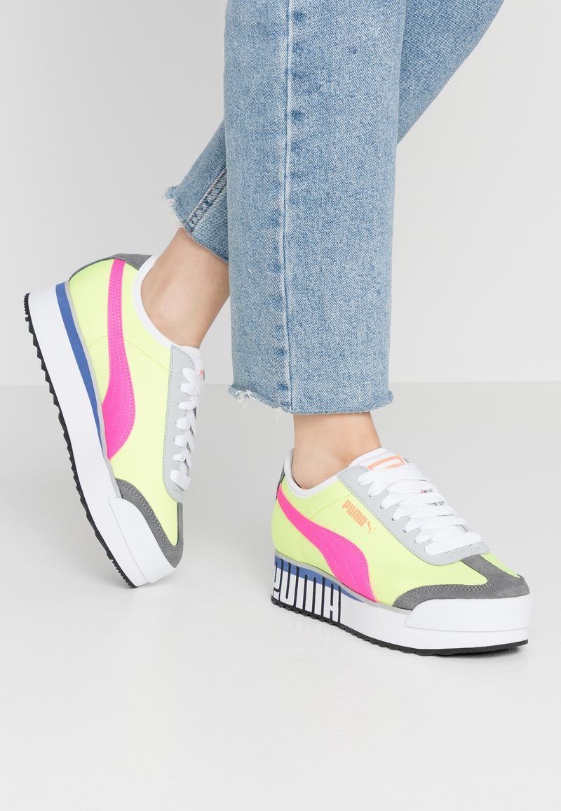 Jambes en jean bleu clair portant des baskets plateforme Puma néon jaune, rose, gris et blanc avec des lacets blancs sur un sol blanc.