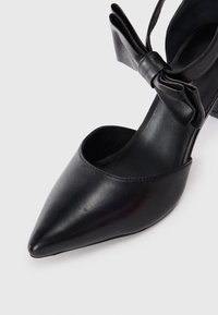 Black leather high-heeled shoe with a pointed toe, open sides, and a decorative bow accent on the ankle strap. Smooth texture and sleek design.