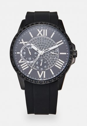 Montre-bracelet Guess noire avec bracelet en silicone noir, chiffres romains, trois sous-cadrans, centre de cadran incrusté de cristaux et lunette ornée de pierres noires.