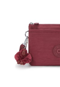 Sacchetto di tessuto bordeaux con chiusura a zip, dotato di un charm a forma di scimmia in gomma e un'etichetta rotonda con logo sulla parte anteriore. Texture liscia, design compatto.