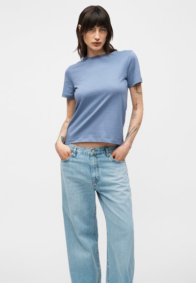 Γυναίκα με σκούρα μαλλιά που φοράει ανοιχτό μπλε κοντομάνικο πουκάμισο και ανοιχτό μπλε τζιν ψηλόμεσα, στέκεται με τα χέρια στις τσέπες μπροστά σε λευκό φόντο.