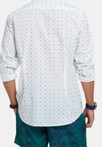 Camisa blanca con pequeños patrones azules, mangas largas arremangadas, dobladillo curvado, textura similar al algodón. Combinada con shorts oscuros de patrón verde.
