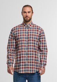 Mann mit Bart, der ein knopfbares, langärmeliges, mehrfarbiges kariertes Hemd und blaue Jeans trägt und vor einem schlichten Hintergrund steht.