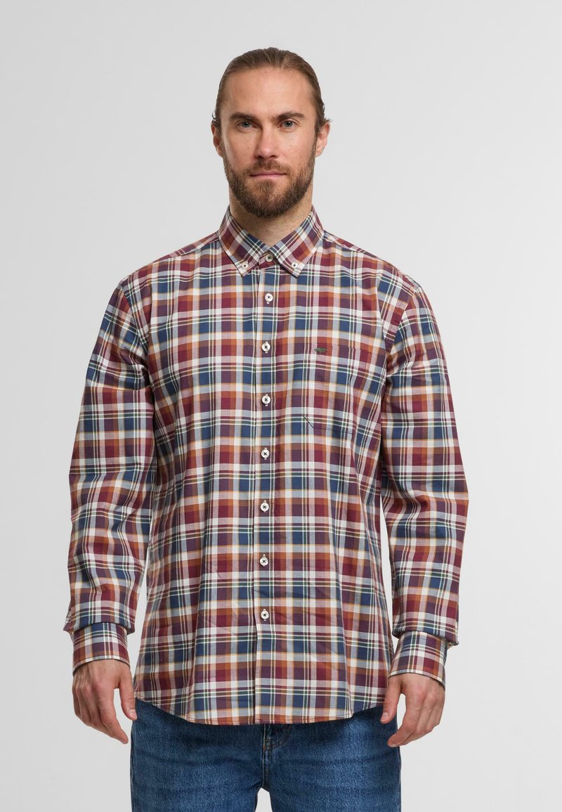 Mann mit Bart, der ein knopfbares, langärmeliges, mehrfarbiges kariertes Hemd und blaue Jeans trägt und vor einem schlichten Hintergrund steht.