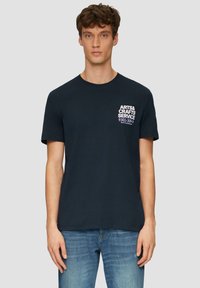 Navyblauwe katoenen T-shirt met korte mouwen, ronde hals en witte tekstgrafiek op het linkerbovenlichaam. Gecombineerd met lichtblauwe spijkerbroek.