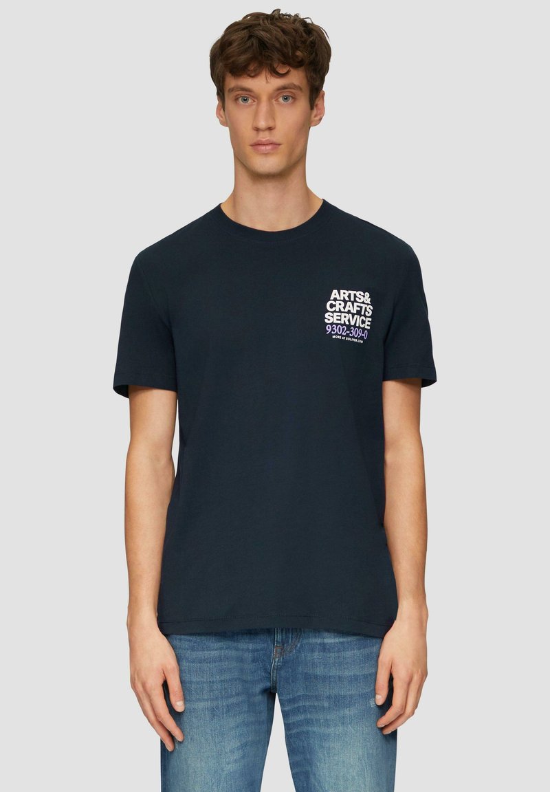 Navyblauwe katoenen T-shirt met korte mouwen, ronde hals en witte tekstgrafiek op het linkerbovenlichaam. Gecombineerd met lichtblauwe spijkerbroek.