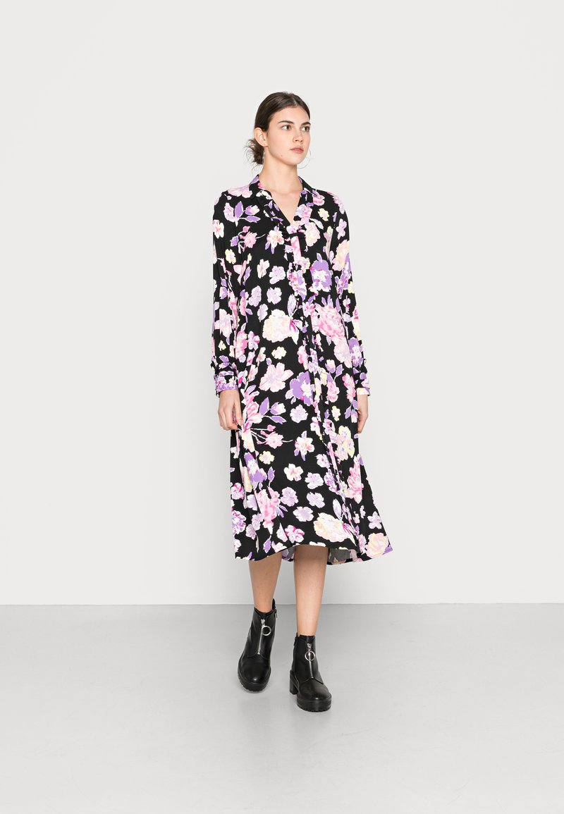Vestido preto floral com padrões de flores roxas e pink, mangas longas, frente com botões e uma saia fluida de comprimento médio. Botas pretas até ao tornozelo.