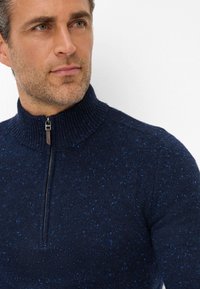 Marineblauer Strickpullover mit hohem Kragen, der einen Viertel-Reißverschluss und kleine Sprenkel in hellblau im Stoff aufweist.