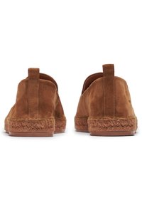 Des chaussures à enfiler en daim marron avec une semelle en jute et un petit tirant à l'arrière. Texture lisse avec une forme arrondie et un design minimal.