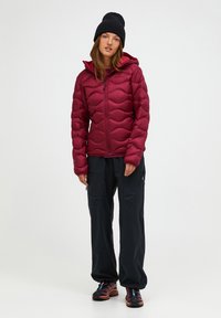 Burgundy puffer jacket with wave pattern, front zipper, and hood; paired with loose black pants and hiking shoes with orange accents.