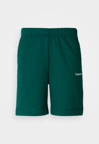 Grønne bomuldsshorts med elastik i taljen, med et logo på den nedre højre side og syede kanter ved benåbningerne.