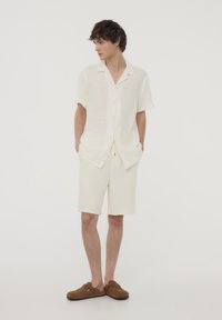 Camicetta a maniche corte con colletto in lino color avorio con una texture sottile, abbinata a pantaloni corti avorio e scarpe slip-on marroni.