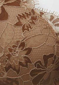 Beige kantstof met bloemmotieven in donkerbruin tonen, met delicate borduurwerk en een zachte, gestructureerde afwerking.