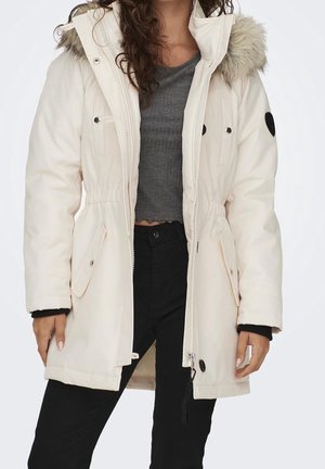 Kobieta ubrana w długi, biały zimowy płaszcz z futrzanym kapturem, pod którym ma szary krótki top i czarne spodnie, stoi ze swobodnie opuszczonymi rękami.