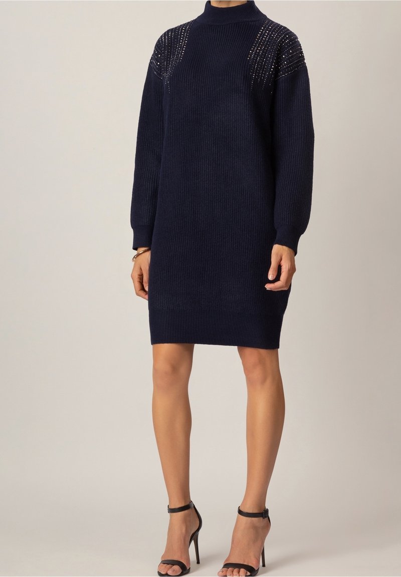 Robe-pull en maille bleu marine avec détails cloutés sur les épaules, manches longues et col montant, portée avec des escarpins noirs à bride à la cheville.