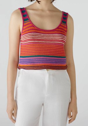 Kvinde iført ærmeløs, flerfarvet stribet strikket cropped top kombineret med højtaljede hvide bukser på en ensfarvet baggrund.