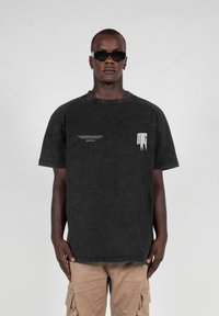 Black denim t-shirt with a round neckline, short sleeves, and white printed accents on the chest and side. Worn with khaki cargo shorts.