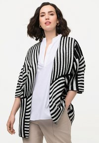 Randig svart och vit cardigan med avslappnad passform, öppen frampart, och ärmar i armbågs-längd, kombinerad med en vit skjorta och beige byxor.