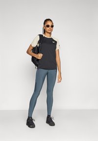 Sort og beige sports-T-shirt, blå leggings med sømdetaljer og sorte sko. Modellen holder en sort rygsæk og har solbriller på.