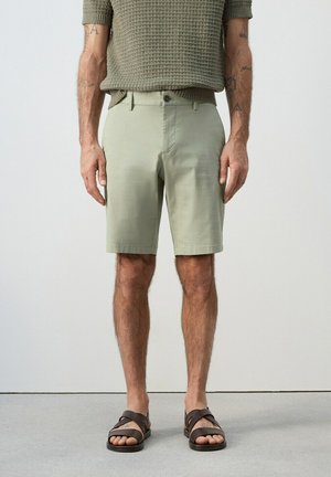 Uomo che indossa pantaloncini verde chiaro al ginocchio, maglione a maniche corte color oliva con trama, e sandali marrone scuro, in piedi davanti a uno sfondo semplice.
