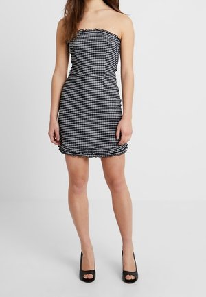 Woman wearing a strapless, black and white checkered mini dress with ruffled edges and black open-toe high heels, standing against a plain background.