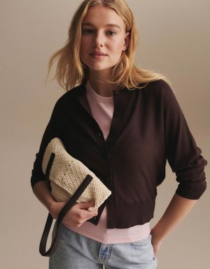 Giovane donna con capelli biondi che indossa un cardigan scuro abbottonato sopra una maglietta rosa, che tiene in mano una clutch lavorata a maglia color crema con una tracolla nera.